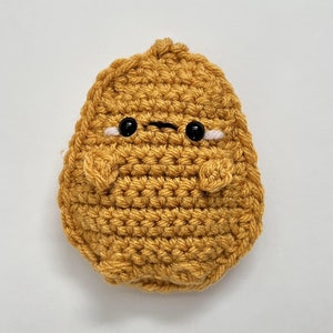 Könnte beinhalten: Ein gehäkeltes Kartoffelspielzeug in gelb-brauner Farbe mit einem einfachen Ausdruck mit schwarzen Augen. Die Kartoffel hat einen leicht mürrischen Blick.