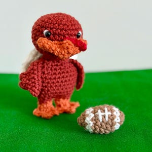 Può includere: Un'anatra all'uncinetto fatta a mano nei toni del rosso, arancione e bianco, accanto a un pallone da football americano marrone e bianco. L'anatra ha un corpo rosso, becco e zampe arancioni e ali bianche. Il pallone è marrone con cuciture bianche.