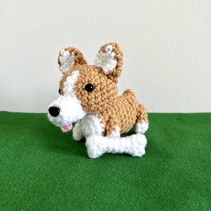 May include: A crocheted stuffed dog toy with brown and white fur. The dog is lying down with its paws outstretched and has a white bone in its mouth.