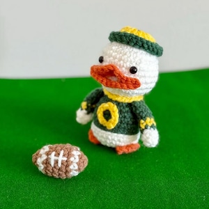 May include: Crocheted duck wearing a green and yellow football uniform, with a matching headband. The duck has an orange beak and is next to a brown and white crocheted football. The duck is on a green surface.