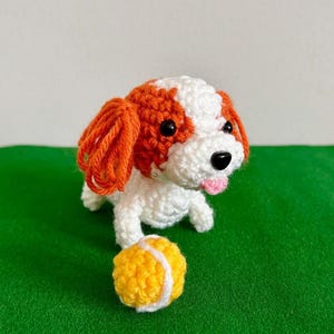 May include: A handmade crochet dog with white and orange fur, black eyes, and a pink tongue. A yellow and white tennis ball sits in front of the dog. The dog is on a green surface.