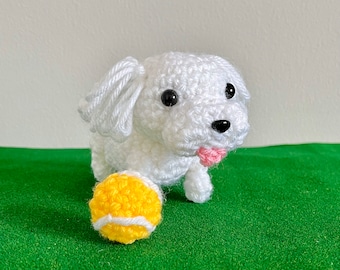 May include: A crocheted white dog with black eyes and a pink tongue, beside a yellow and white crocheted tennis ball. The dog has floppy ears and is on a green surface.