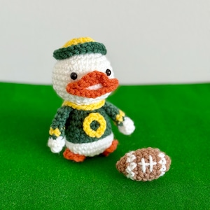May include: A handmade crochet duck wearing a green and yellow football uniform and a matching hat. The duck has an orange beak and is next to a brown and white football. The duck is on a green surface.