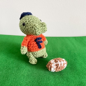 May include: A handmade crochet alligator doll wearing an orange shirt with a blue "F" and a blue hat, next to a brown and white crochet football. The doll is green and the football is brown and white. The background is green.