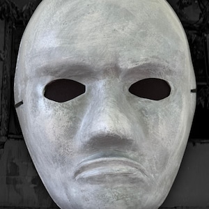 May include: A full-face mask with a textured, weathered light gray finish resembling stone. The mask features oval eye cutouts with dark interiors, a prominent nose, and a straight, neutral mouth line. Text at the bottom reads THE STONEFACE.