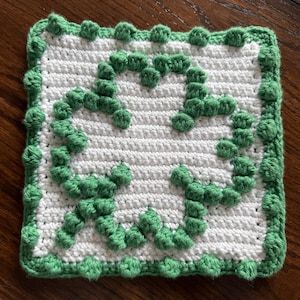 May include: A square, handmade crochet potholder with a white base and a green shamrock design. The shamrock is made with raised, textured stitches. The border and the shamrock's details are in a vibrant green color.