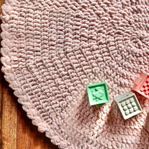 May include: A pink crocheted rug with a textured surface. Three colorful silicone baby teethers are on the rug. One teether is green with a fish design, one is gray with a bumpy surface, and one is pink with a grid pattern.