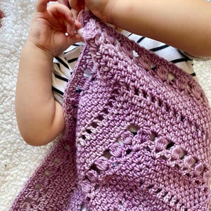 May include: A close-up of a purple crocheted baby blanket with a delicate pattern. The blanket is soft and cosy, perfect for a newborn.