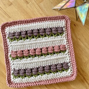 Könnte beinhalten: Ein quadratischer, gehäkelter Gegenstand mit Reihen von lila und rosa tulpenartigen Blumen auf weißem Hintergrund, umrahmt von einem rosa Rand. Die Blumen haben grüne Stiele. Es ist wahrscheinlich ein dekoratives Stück oder eine kleine Decke.