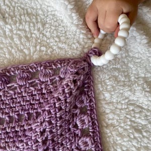 May include: A purple crocheted baby blanket with a white teething ring attached to one corner.