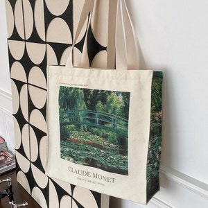 Puede incluir: Una bolsa de lona blanca con una imagen de un puente verde y un estanque de nenúfares. La bolsa tiene un estampado de Claude Monet con el texto "The Water Lily Pond".