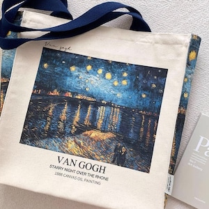 Op de afbeelding: Een crèmekleurige canvas draagtas met een marineblauwe band, met Van Goghs "Sterrennacht over de Rhône". De tas toont de naam van de kunstenaar en de beschrijving van het schilderij. Een praktische accessoire.