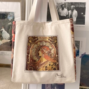 Puede incluir: Una bolsa de lona blanca con una impresión de arte vintage colorida que presenta el perfil de una mujer y el texto "La Plume" y "Mucha". La bolsa tiene tela con estampado floral rojo y dorado en los lados.