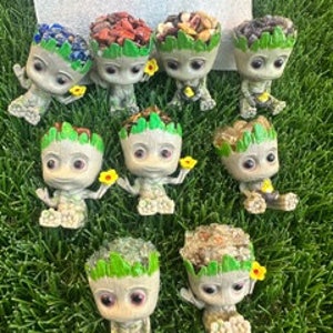 May include: Eight small, gray, resin figurines of a character from the Guardians of the Galaxy movie, Groot. Each figurine is sitting with its legs crossed and holding a small yellow flower. Each figurine has a different type of greenery and natural elements on its head, such as blue and purple flowers, red berries, nuts, and brown seeds.