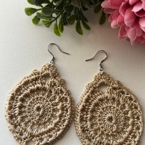 May include: A pair of beige crocheted earrings with a delicate floral design. The earrings are made with a fine yarn and have a simple, elegant style.