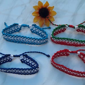 May include: Four handmade friendship bracelets in blue, red, and green with white accents. The bracelets are made of woven thread and are displayed with a yellow sunflower.