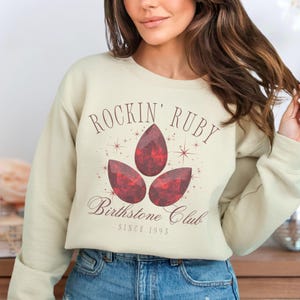 May include: A cream colored sweatshirt with the text "Rockin' Ruby Birthstone Club Since 1995" and a graphic of three red ruby gemstones.