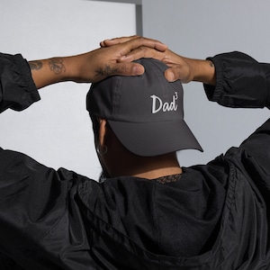 May include: A dark gray baseball cap with the word "Dad" embroidered in white on the front. The cap has a curved brim and a mesh back.