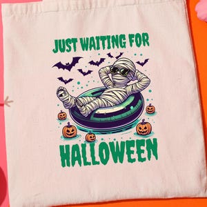 May include: A cream tote bag with a Halloween design. It features a mummy relaxing on a pool float, surrounded by pumpkins and bats, with the text "JUST WAITING FOR HALLOWEEN". The background is pink and orange. The bag is a seasonal item.