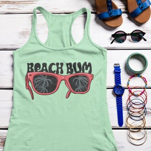 May include: A light green racerback tank top featuring a graphic of red sunglasses with palm tree silhouettes in the lenses and the text "BEACH BUM" in a dark, wavy font. The design is centered on the front of the garment.