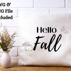 Op de afbeelding: Een wit kussen met zwarte tekst die "Hello Fall" zegt met een kleine pompoen en bladeren in de hoek.