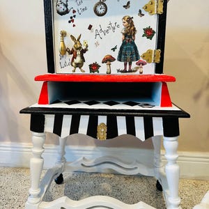 May include: A whimsical, hand-painted side table with an Alice in Wonderland theme. The table features a black and white striped top, white legs, and a red shelf. The top section has a decorative panel with illustrations and the text "Adventure".
