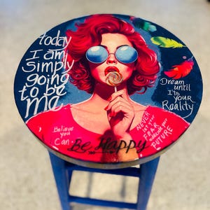 May include: A round wooden stool with a blue base and a top featuring a colorful illustration of a person with red hair, blue sunglasses, and a lollipop. The text reads: Today I am simply going to be me, Dream until it's your reality, Believe you can, Be happy, and Never let your fear decide your future.