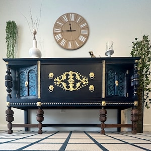 May include: A black and gold vintage sideboard with ornate details, featuring two doors and three drawers. The sideboard has a dark wood base and gold accents.