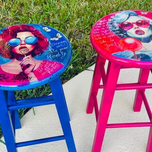May include: Two colorful bar stools. One is blue with a round seat featuring a woman with red hair, sunglasses, and a lollipop, with text. The other is pink with a similar design, but with a different woman and text. Both have a three-legged design.