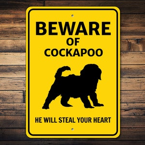 Puede incluir: Señal de advertencia amarilla con texto negro que dice "Cuidado con el Cockapoo. Te robará el corazón." Una silueta negra de un perro Cockapoo está en el centro del cartel.