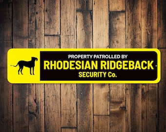 Puede incluir: Un letrero amarillo y negro con el texto "PROPERTY PATROLLED BY RHODESIAN RIDGEBACK SECURITY CO.". Una silueta negra de un perro Rhodesian Ridgeback está en el lado izquierdo del letrero.