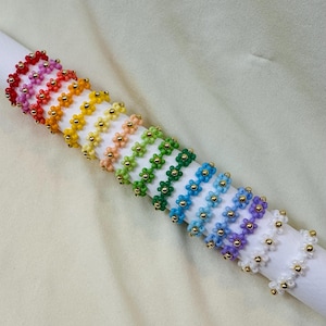 May include: A set of 12 colorful beaded rings with flower designs. The rings are arranged in a rainbow pattern, with each ring featuring a different color.