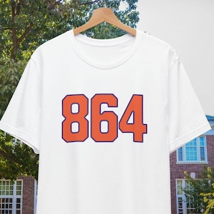 Può includere: Maglietta bianca con il numero 864 stampato in arancione con un contorno blu scuro.