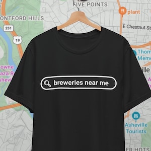 Può includere: Una maglietta nera con una grafica a barra di ricerca bianca che dice "breweries near me" su una maglietta nera. La maglietta è appesa a un appendiabiti davanti a una mappa di Asheville, Carolina del Nord.