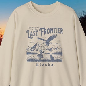 May include: A cream-coloured sweatshirt with a blue graphic print of a mountain scene with a bald eagle flying overhead. The text "Welcome to the Last Frontier Alaska" is printed on the sweatshirt.
