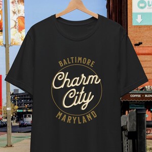 May include: Black t-shirt with a gold circle graphic that says "Baltimore Charm City Maryland".