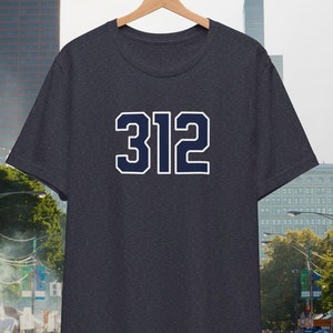 May include: A dark heather grey t-shirt with a navy blue 312 printed on the front. The t-shirt is hanging on a wooden hanger against a city backdrop.