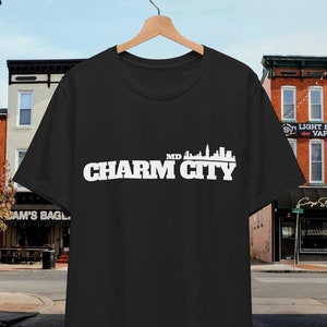 Puede incluir: Camiseta negra con un gráfico blanco del horizonte de Baltimore y el texto "Charm City MD".