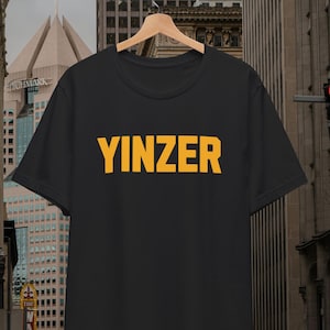 May include: Black t-shirt with the word "YINZER" printed in yellow block letters. The shirt is hanging on a wooden hanger against a blurred city background.