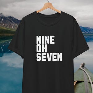 May include: Black t-shirt with the text "NINE OH SEVEN" in bold white letters. The shirt is hanging on a wooden hanger against a backdrop of a lake and mountains. The shirt is a classic crew neck style.