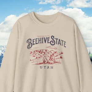 Könnte beinhalten: Cremefarbenes Sweatshirt mit der Aufschrift "Welcome to The Beehive State" über einer Illustration von Landschaft und Figuren. Das Wort "UTAH" ist darunter aufgedruckt. Das Sweatshirt hat einen Rundhalsausschnitt und lange Ärmel.