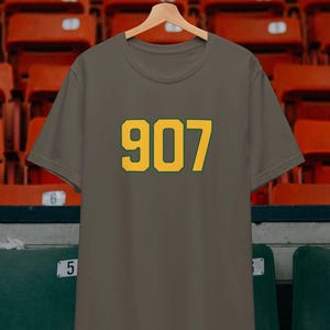 May include: An olive green t-shirt with the number "907" printed in yellow with a green outline. The shirt is hanging on a wooden hanger. The background shows stadium seating.