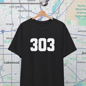 May include: Black t-shirt with the number "303" in bold white font. The shirt is hanging on a wooden hanger against a map background. The map shows the Denver, Colorado area.