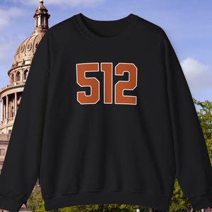 May include: Black sweatshirt with the number "512" in orange block letters. The sweatshirt is a crew neck style. The background features a building with a dome and a grassy area.