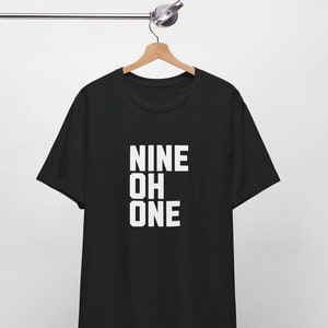 May include: Black t-shirt with the white text "NINE OH ONE" printed vertically. The shirt is hanging on a wooden hanger against a white background. The t-shirt is made of a soft material.