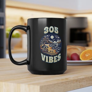 Puede incluir: Taza de cerámica negra con un gráfico blanco de una escena de playa con palmeras, una luna y el texto "305 VIBES".