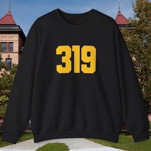 Op de afbeelding: Zwart sweatshirt met het nummer "319" in gouden blokletters. Het sweatshirt heeft een ronde hals. De achtergrond toont een gebouw met een rood dak en een blauwe lucht.
