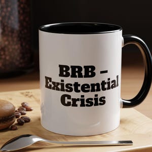 Puede incluir: Taza de cerámica blanca con borde y asa negros. La taza tiene texto negro que dice "BRB - Existential Crisis".