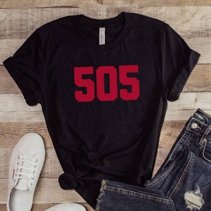 May include: Black t-shirt with the number "505" in red block letters. The shirt is paired with blue denim jeans and white sneakers. The items are arranged on a wooden surface.