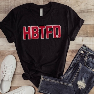 May include: Black t-shirt with the text "HBTFD" in red letters. The shirt is paired with blue jeans and white sneakers.